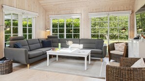 Un salon chaleureux avec un canapé d’angle gris, une table basse blanche, des fauteuils en osier et de grandes fenêtres donnant sur un paysage verdoyant.