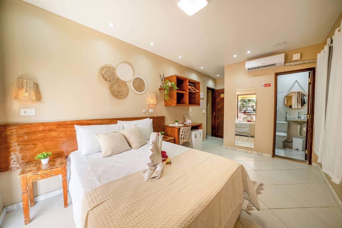 Une chambre d’hôtel avec une tête de lit en bois, un lit recouvert de draps blancs, une table de chevet avec une lampe, une petite plante et un miroir au mur.