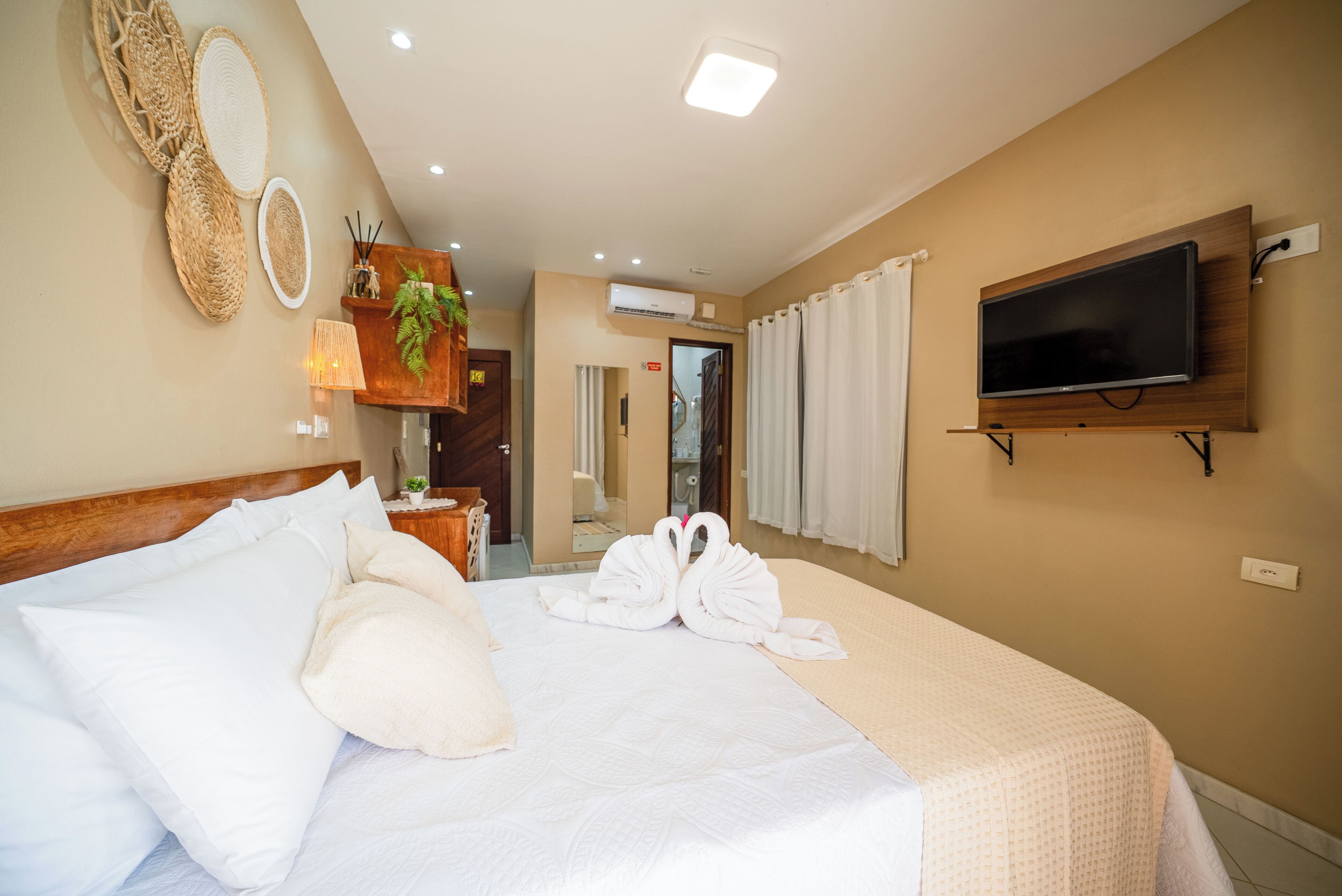A hotel room with a large bed, a flat-screen TV mounted on the wall, and a small desk with a lamp and decorative items.