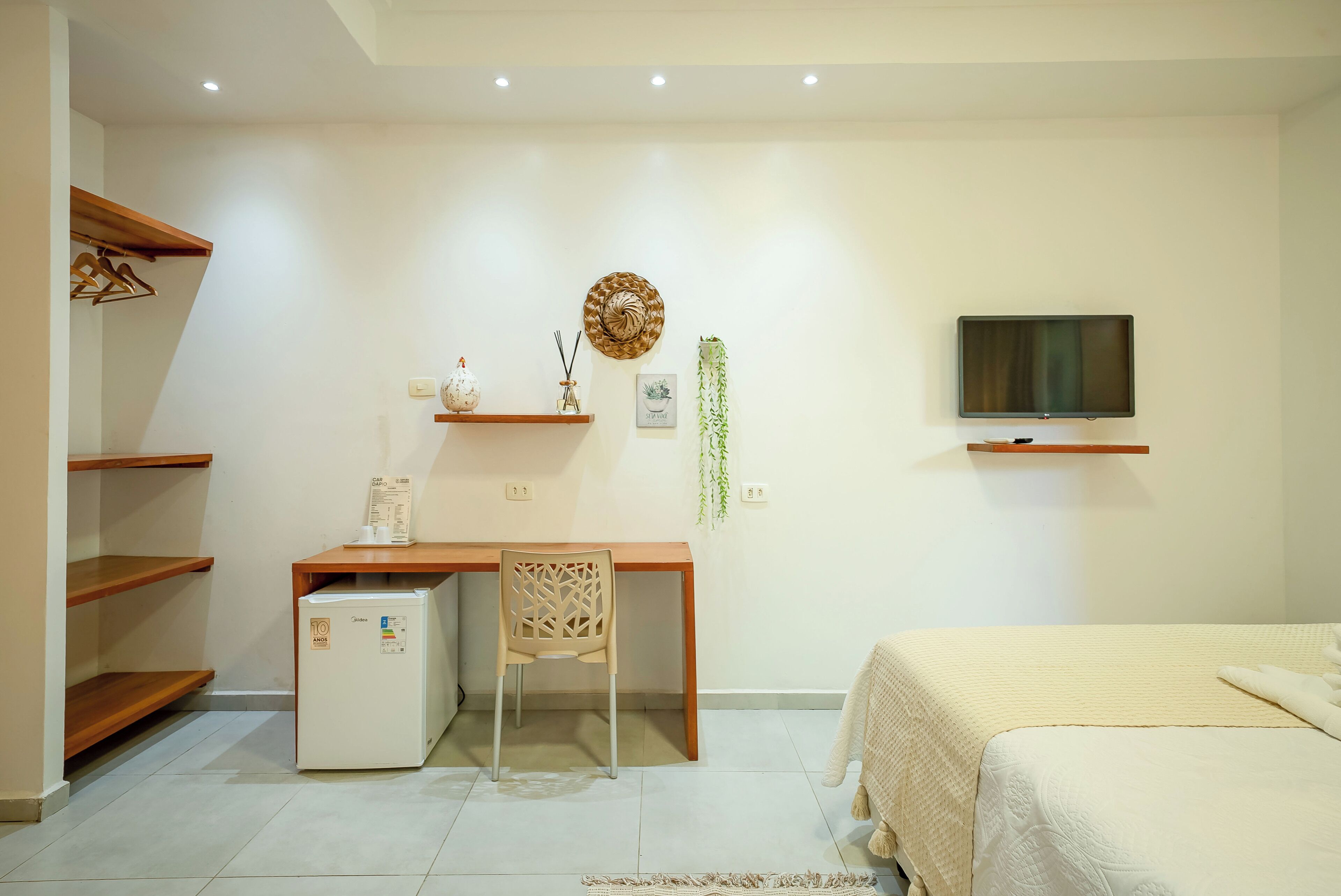 A bedroom with a bed, a desk with a chair, a small refrigerator, and a television on the wall.
