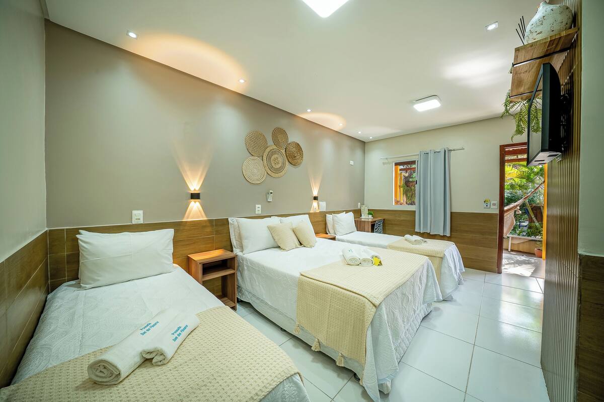 Une chambre d’hôtel avec trois lits, une tête de lit en bois, un téléviseur fixé au mur et une fenêtre avec des rideaux.