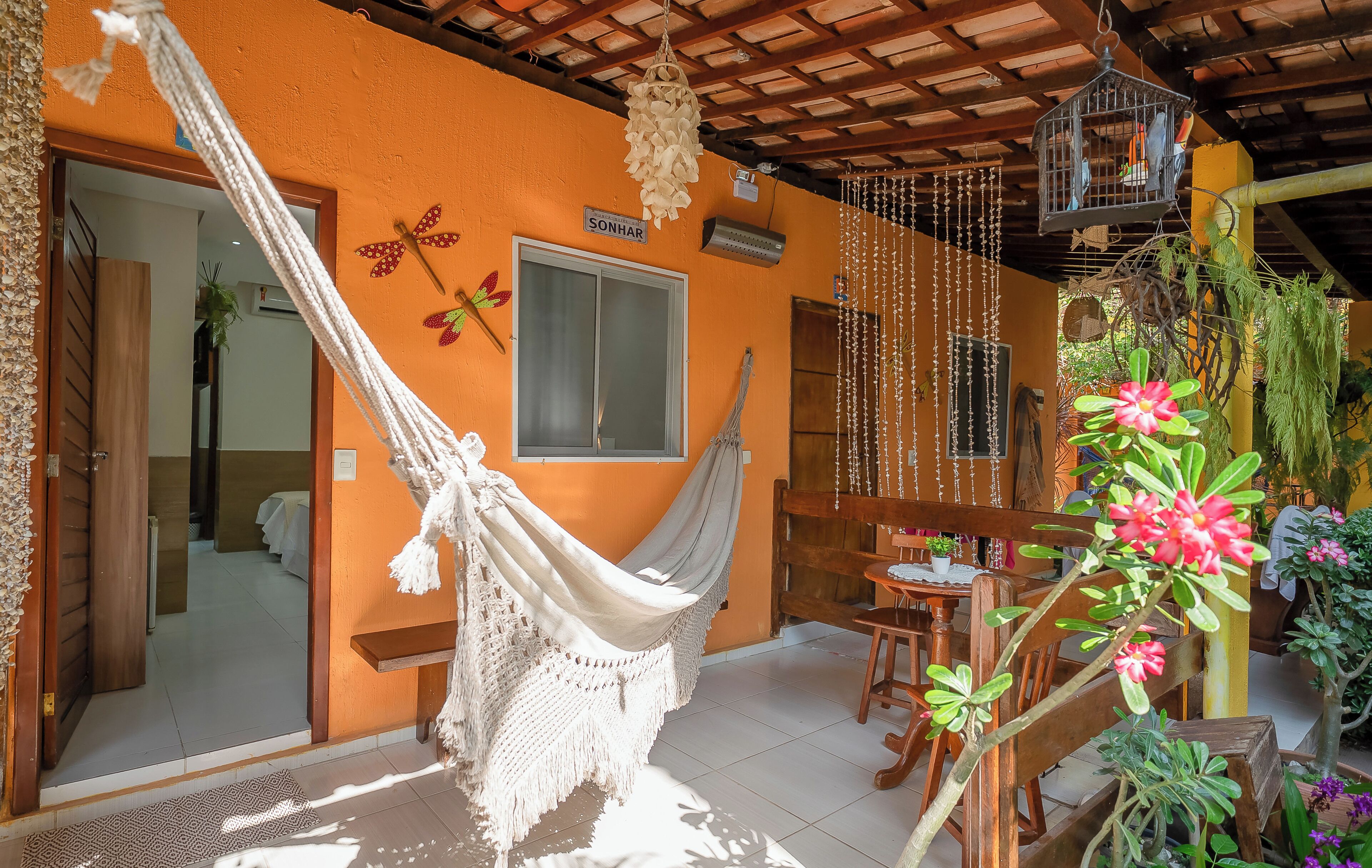 A hammock hanging in front of an orange wall with a wooden ceiling, a wooden chair, and a table with a plant.