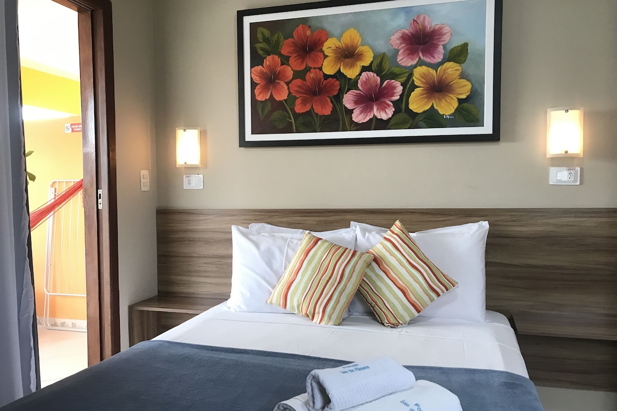 A hotel room with a bed, pillows, a painting, wall sconces, and a view of the outside.