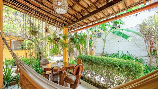 A hammock with wooden chairs and a table set up under a wooden pergola with hanging plants. - Pousada Som dos Passaros (Ipojuca)