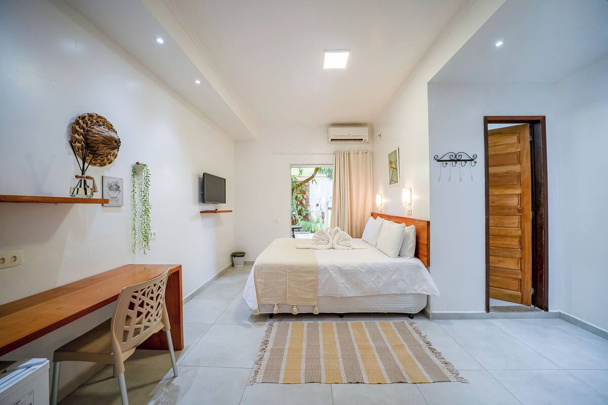 Une chambre avec un lit, une table de chevet et un bureau avec une chaise. Il y a un téléviseur accroché au mur, une fenêtre avec des rideaux et une porte.