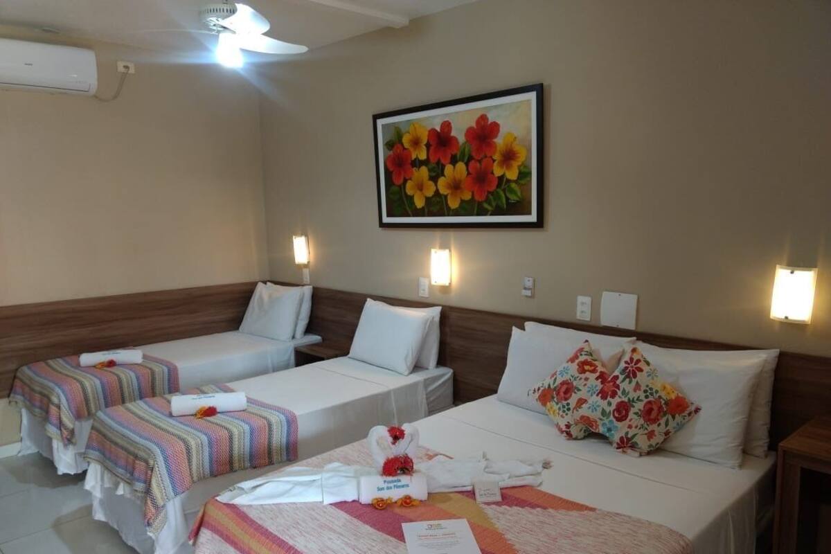 Une chambre d’hôtel avec trois lits, un ventilateur de plafond, un appareil de climatisation et un tableau représentant des fleurs accroché au mur.