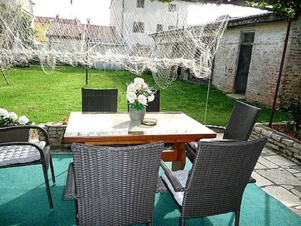 An outdoor patio with a table and chairs, a white decorative net, and a grassy area with a building in the background.