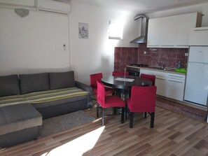 Een knus woongedeelte met een bank, een eettafel, stoelen, een keuken en een raam.