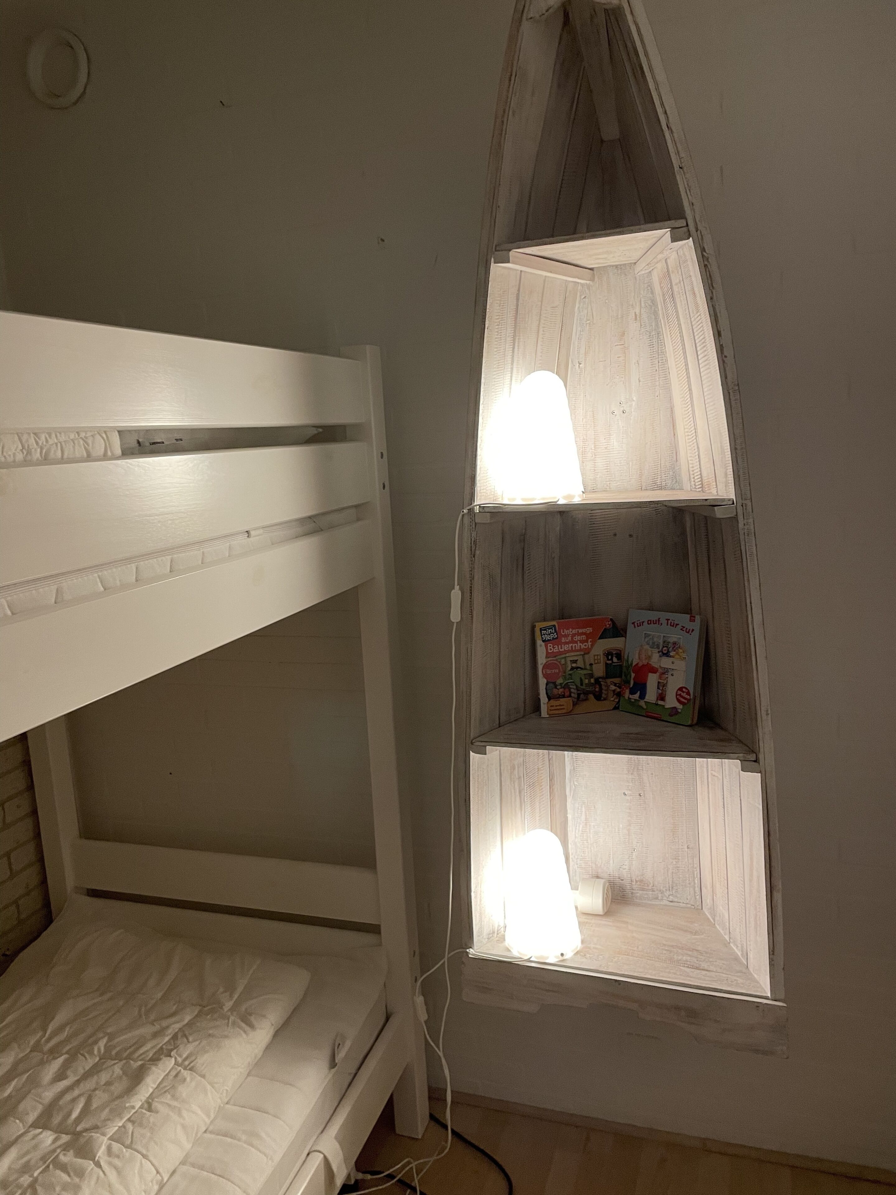 A bunk bed with a shelf unit attached to the wall, containing children's books and a small window with light fixtures.