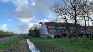 A two-story building with a red roof and a white tower, adjacent to a grassy area with a small stream. - Cozy and friendly vacation apartment in a quiet location. (Bruinisse)
