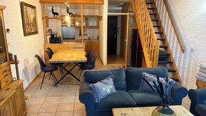 A cozy living room with a dark blue sofa, a wooden coffee table, and a dining area with a wooden table and chairs. A staircase with wooden railings leads to an upper level. - Cozy and friendly vacation apartment in a quiet location. (Bruinisse)