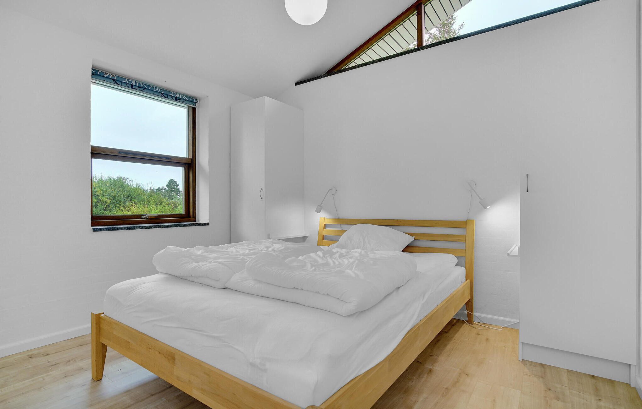 A minimalist bedroom with a wooden bed, white bedding, a large window with a view, and a sloped ceiling.