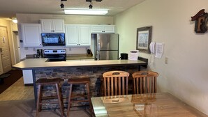 A kitchen with white cabinets, a stainless steel refrigerator, a built-in oven, and a stone backsplash. There is a wooden dining table with four chairs. - Ski In Ski Out Condo (Whitefish)