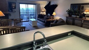 A modern kitchen with a central island, a dining area with chairs, a sofa, and a balcony with a view. - Ski In Ski Out Condo (Whitefish)