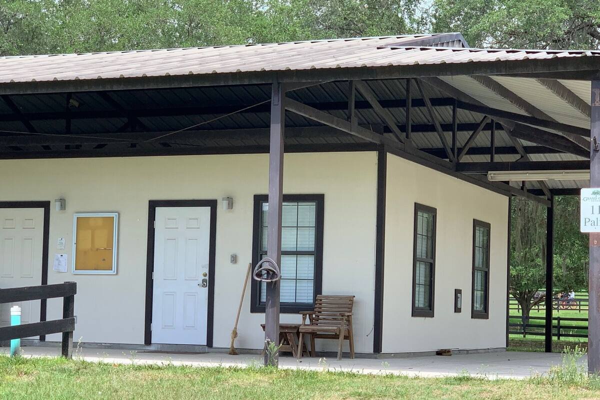 A small, single-story building with a metal roof and a porch. There are two doors, one with a window and one with a solid door. A bench is placed on the porch, and a broom leans against the wall.
