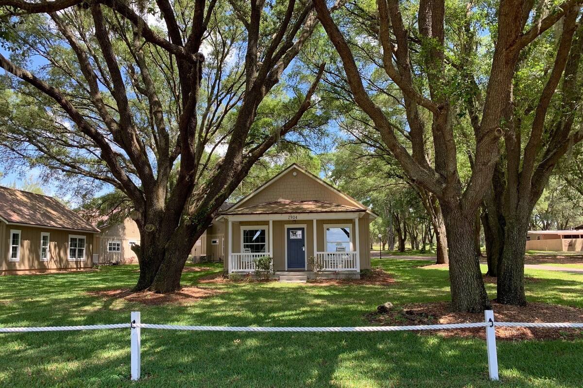 A small, single-story house with a porch, surrounded by large trees.