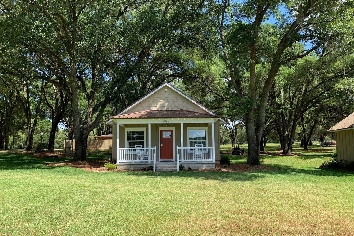 A small, single-story house with a red door and white porch, surrounded by trees.