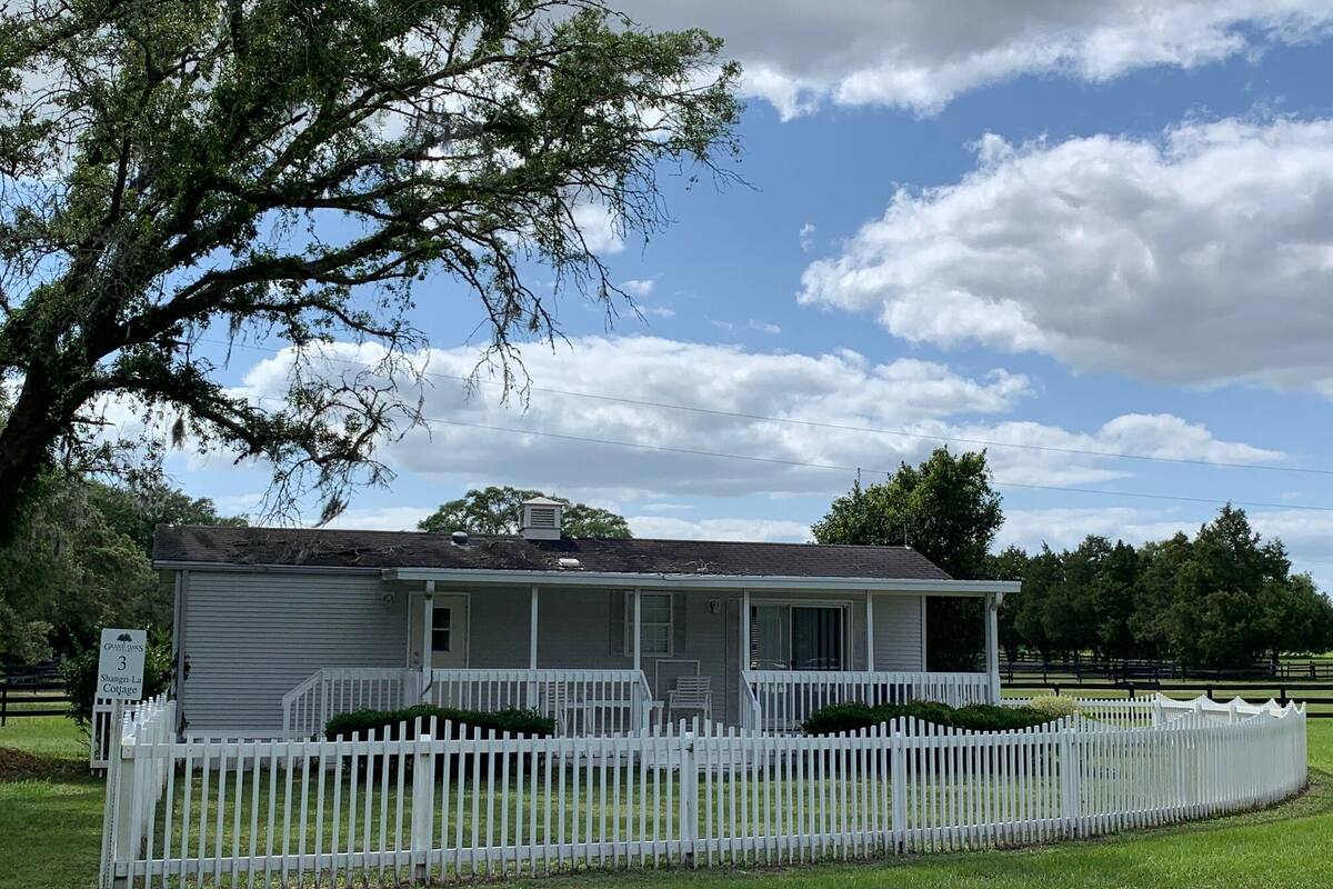 A small, single-story cottage with a white picket fence, a covered porch, and a large tree to the left.