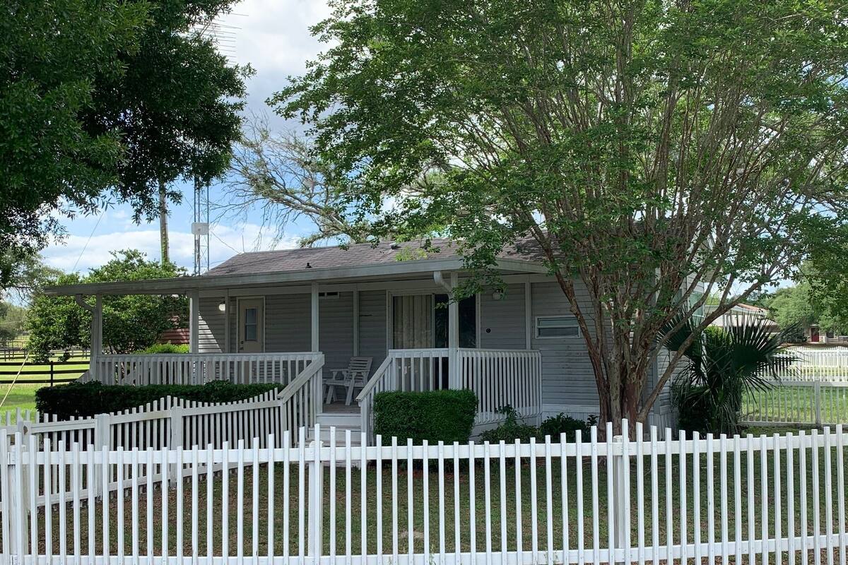 A single-story house with a porch, a white picket fence, and a tree in front.