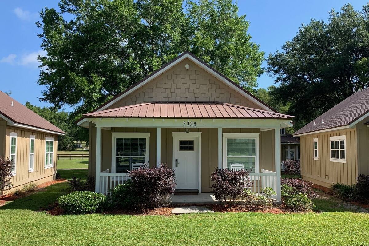 A small, single-story house with a beige exterior and a brown roof, featuring a front porch with a white railing and a small porch area.