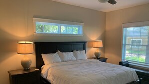 A bedroom with a large bed, two bedside tables, a ceiling fan, a window with blinds, and a view of greenery outside. - Grand Oaks Resort (Lady Lake)