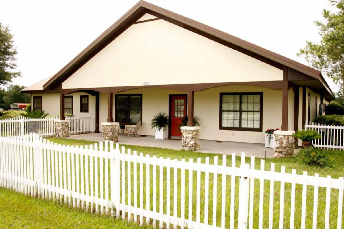 A single-story house with a beige facade, a brown roof, and a white picket fence.