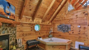 A log cabin interior with a stone fireplace, a wooden bathtub, and a stone wall with a window.
