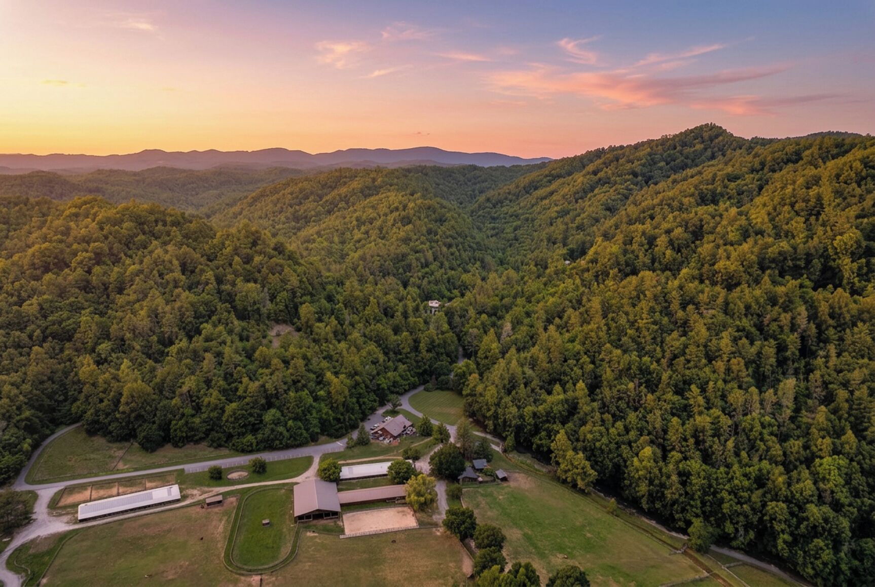 An aerial view of a rural area with dense forests, a winding road, and several buildings including a large warehouse and smaller structures.