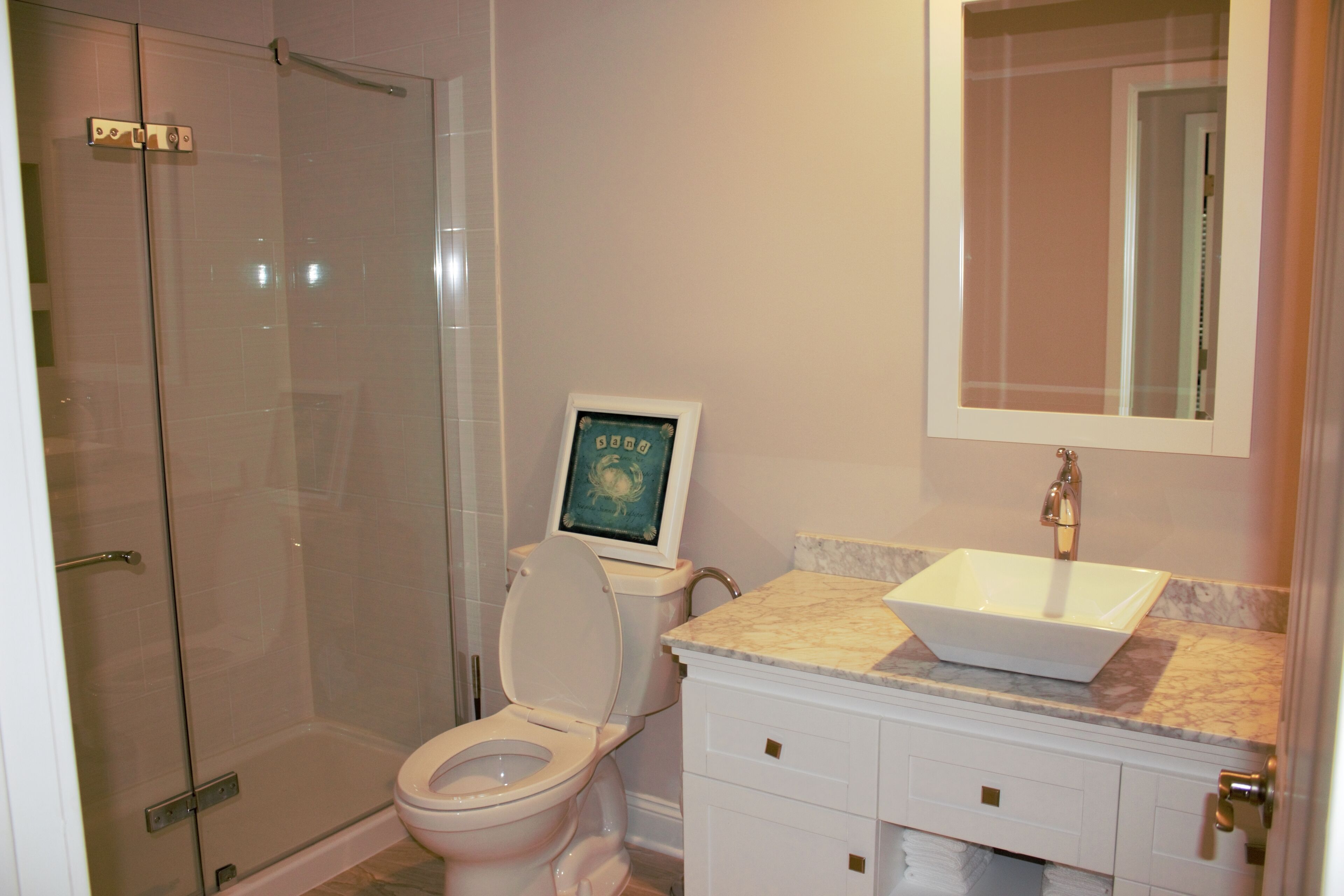 Ein Badezimmer mit Duschkabine aus Glas, einer Toilette mit gerahmtem Bild und einem Waschbecken mit Marmorplatte.