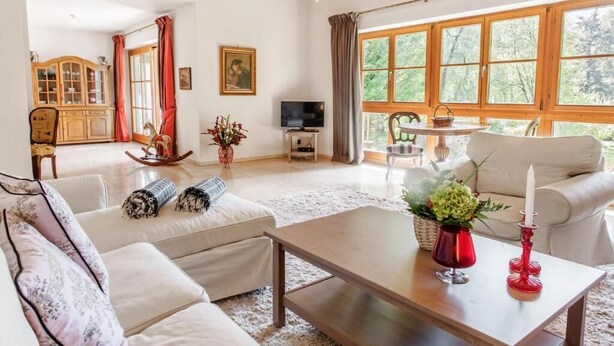 Amplia sala de estar con techo alto, vigas de madera, un ventanal con vistas a árboles, un televisor y una zona de estar acogedora con un sofá y sillones.