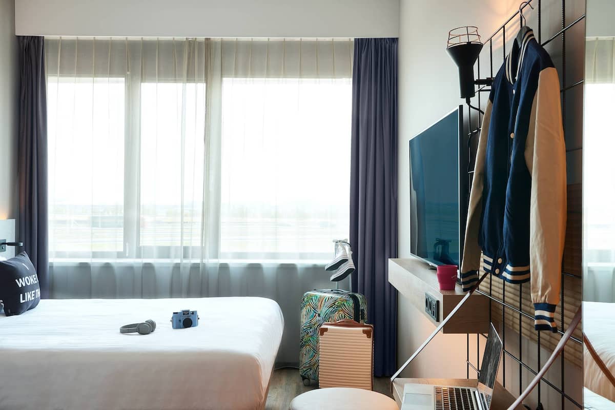 Ein Hotelzimmer mit zwei Betten, einem Fernseher, einem Schreibtisch mit einem Laptop, einem Koffer und einem Kleiderständer mit Jacken.