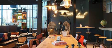 A modern cafe with a long wooden table, wicker chairs, and hanging pendant lights.