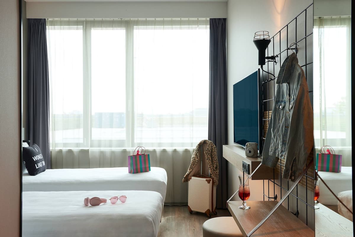 Ein Hotelzimmer mit zwei Betten, einem Fernseher, einem Kleiderständer und einem fensterbepflanzten Fenster.