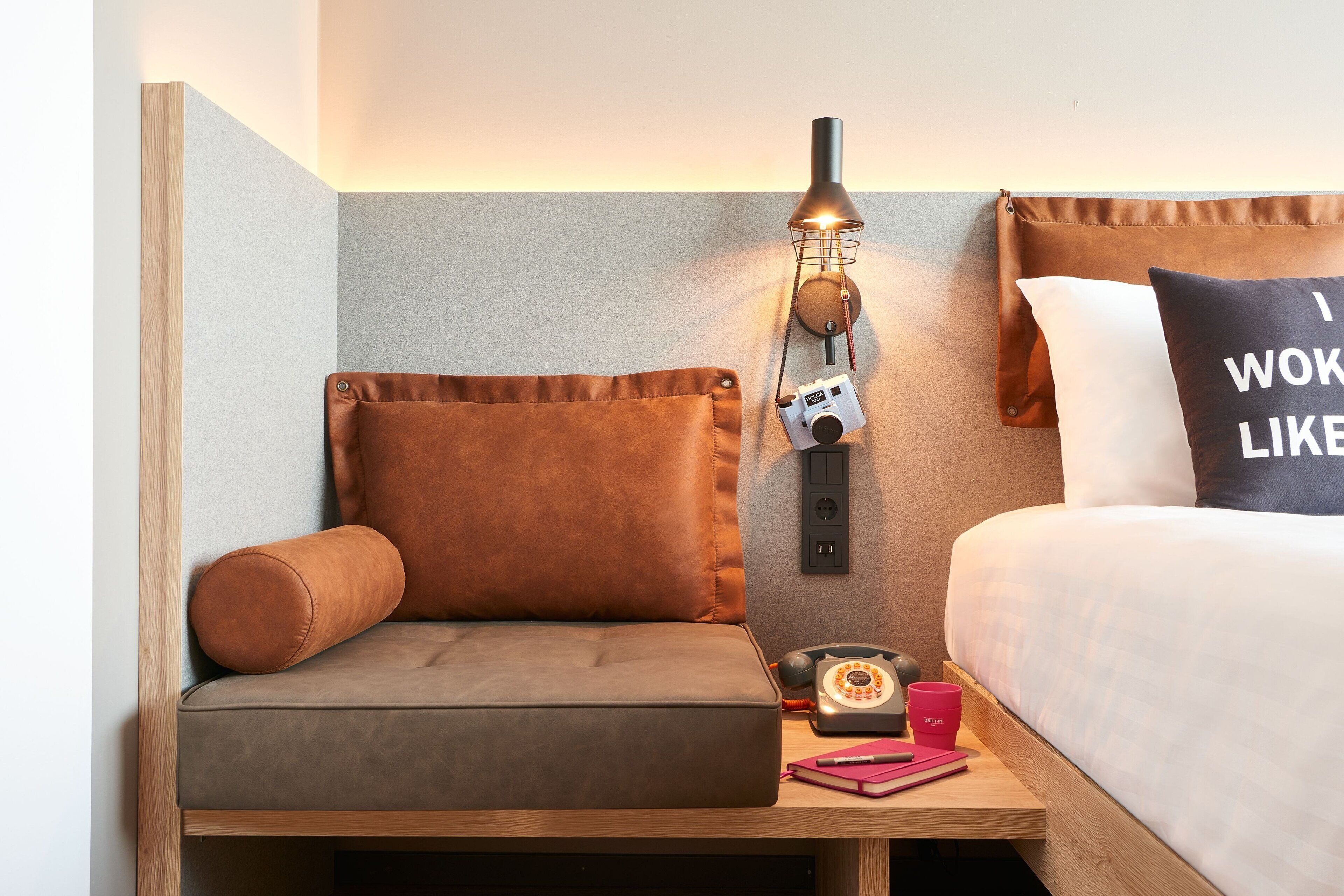 Een gezellige hotelkamer met een houten nachtkastje, een zithoekje met een kussen, een klassieke telefoon en een wandlamp.