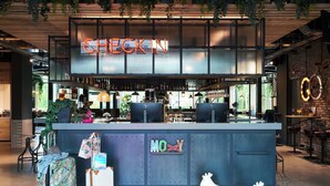 Un espace bar moderne avec un panneau néon indiquant « CHECKIN », un comptoir sur lequel est posé un panneau « NON », et des bagages sur le sol.