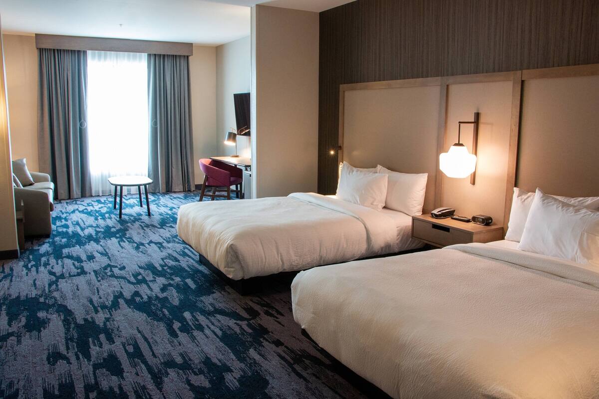 Een hotelkamer met twee bedden, een bureau met stoel, een lamp en een groot raam met gordijnen.