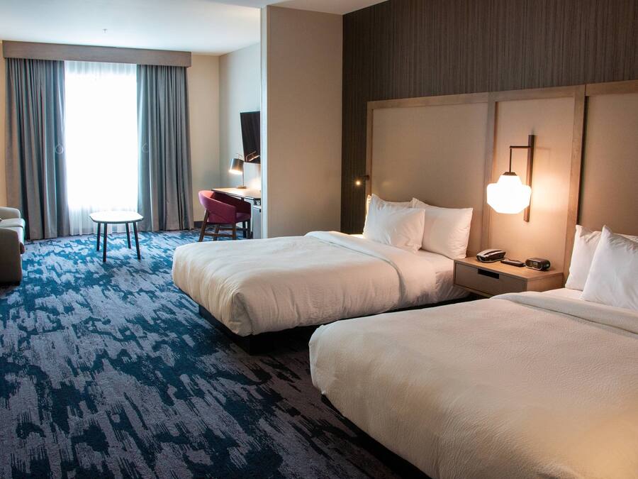 Una habitación de hotel con dos camas, un escritorio con una silla, una lámpara y un ventanal con cortinas.