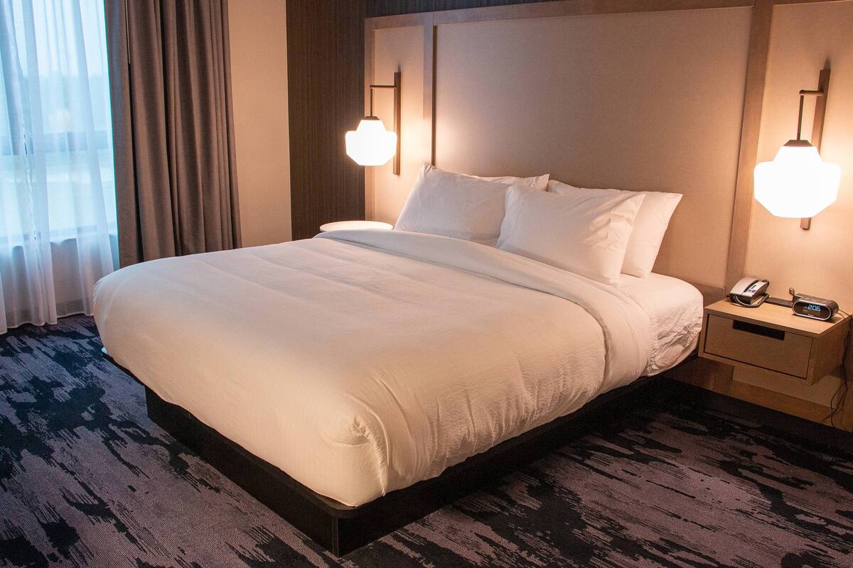 Een net opgemaakt bed met witte lakens, een nachtkastje met een telefoon en een lamp, en een kamerbreed tapijt met een patroon.