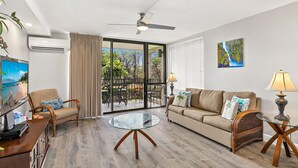 A spacious living room with a glass coffee table, a sofa, a chair, and a desk with a computer. There is a ceiling fan, a window with blinds, and a painting on the wall. - Kamaole Sands 4-213 - Beautiful Modern Renovation - Bright & Open - Split AC (Kihei)