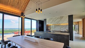 Une cuisine moderne avec un pare-feu en marbre, un coin repas avec une table en bois et des chaises noires, et une vue sur la campagne grâce à de grandes fenêtres.