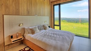 Une chambre à coucher avec un grand lit, une tête de lit en bois, des tables de chevet et une grande fenêtre donnant sur un paysage verdoyant.