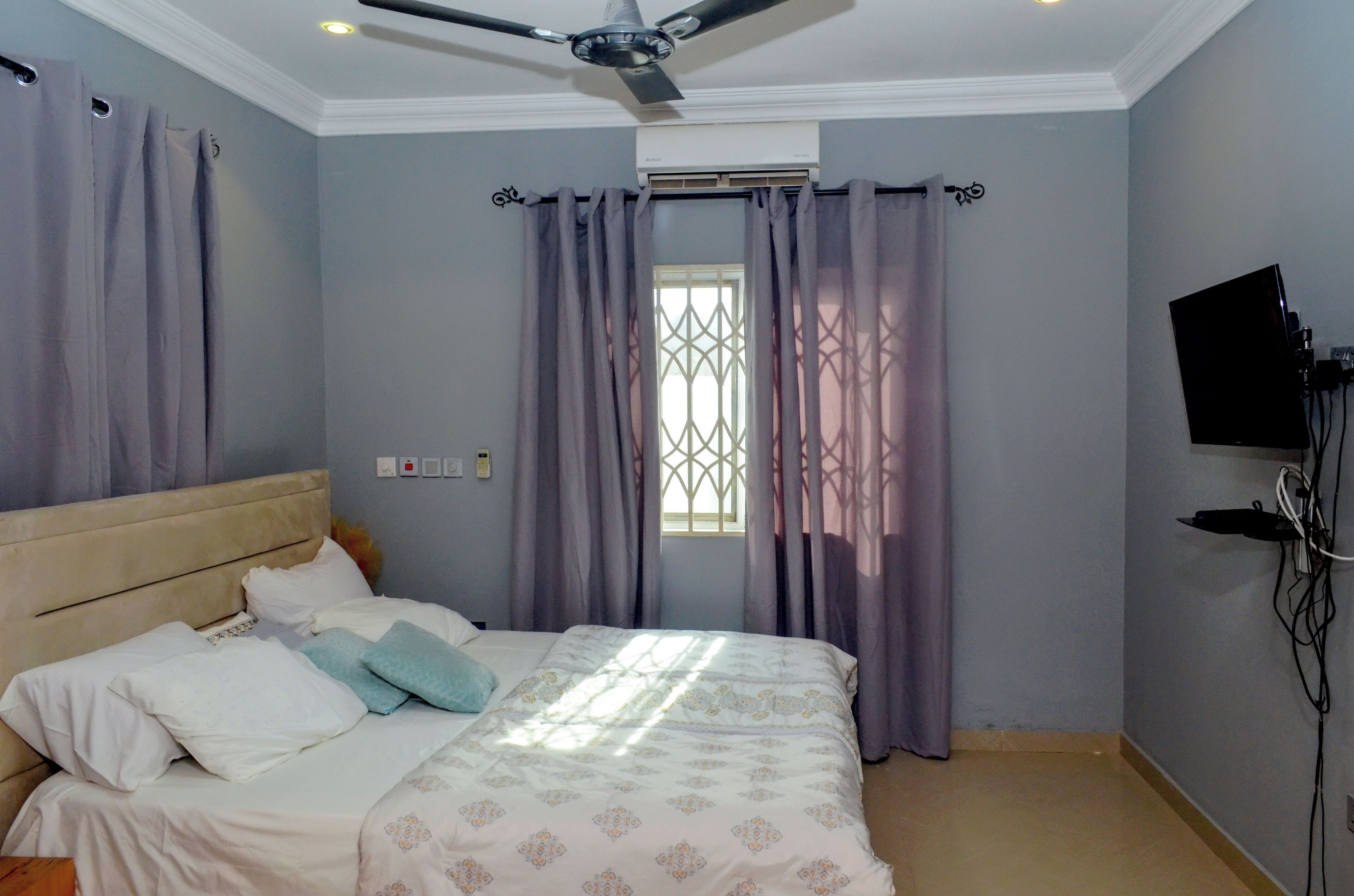 Une chambre à coucher avec un lit, des rideaux, un ventilateur de plafond, une fenêtre et un téléviseur fixé au mur.