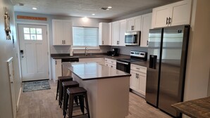 Una cocina moderna con armarios blancos, una isla central con taburetes, electrodomésticos de acero inoxidable y un ventanal con persianas.