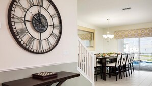 Un comedor con un reloj de pared grande, una mesa de comedor con sillas, y una escalera que conduce a un nivel superior.