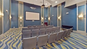 Un salón de conferencias con filas de sillas, una pantalla blanca y lámparas fijadas a la pared.