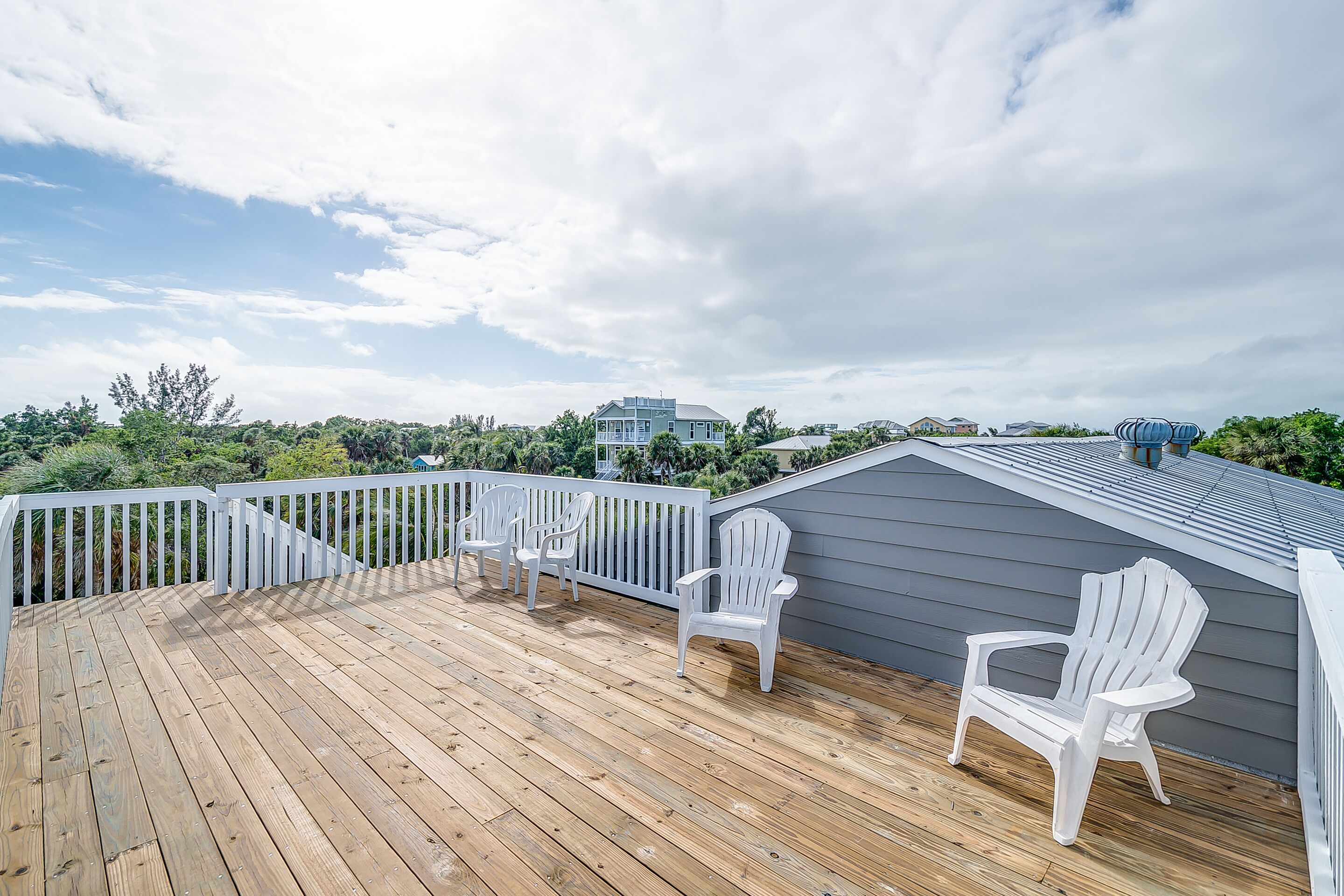 A wooden deck with white Adirondack chairs, a gray building in the background, and a white fence.