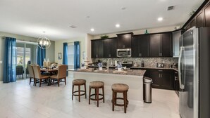 A modern kitchen with dark cabinets, a stainless steel refrigerator, a microwave, and a granite countertop. There is a dining area with a table and chairs, and a trash can is visible.