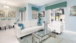 A modern living room with a white sofa, a glass coffee table, and a dining area with white chairs. There are framed artworks on the walls and a large window with blinds. - 9077dd - NON Renting 2-15-21 the Retreat at Championsgate (Davenport)