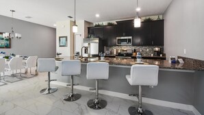 A modern kitchen with dark cabinets, a granite countertop, and a stainless steel refrigerator. There are four white bar stools and a chandelier.