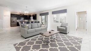 A modern living room with a grey sofa, a coffee table, and a grey armchair. There is a kitchen in the background with dark cabinets and a microwave. - 1668mvd - Non-renting 6-12-2022 The Retreat at Championsgate (Davenport)